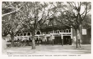 Black and white photograph of a long wooden buildings with tables set up outside the front door…