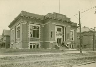 Black and white picture of public library building facing onto a dirt street with single lamppo ...