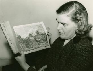 Black and white picture of woman holding a book open to a historical illustration of some build ...