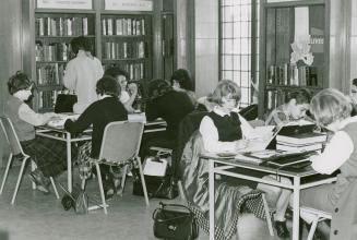 Students seated at tables studying in a library with shelves and window in the background. 