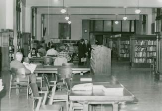 Picture of tables and chairs and people studying in a library room with rows of shelves of book ...
