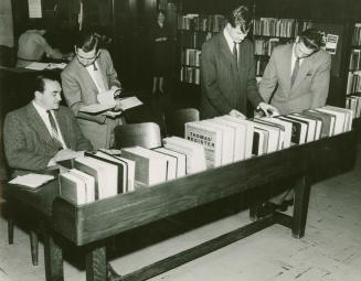 Three men in suits stand looking at a row of business directories and one man is consulting wit ...