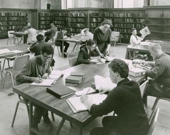 People sit at large tables studying in a large room lined with book shelves and the librarian s ...