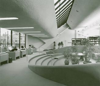 Interior of a library with sunken reading area containing plants and study tables and people si ...