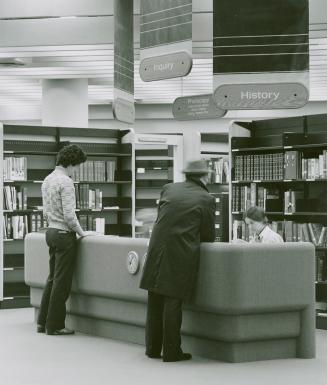 Two men stand at a reference desk in the History Department with the librarian seated behind th ...
