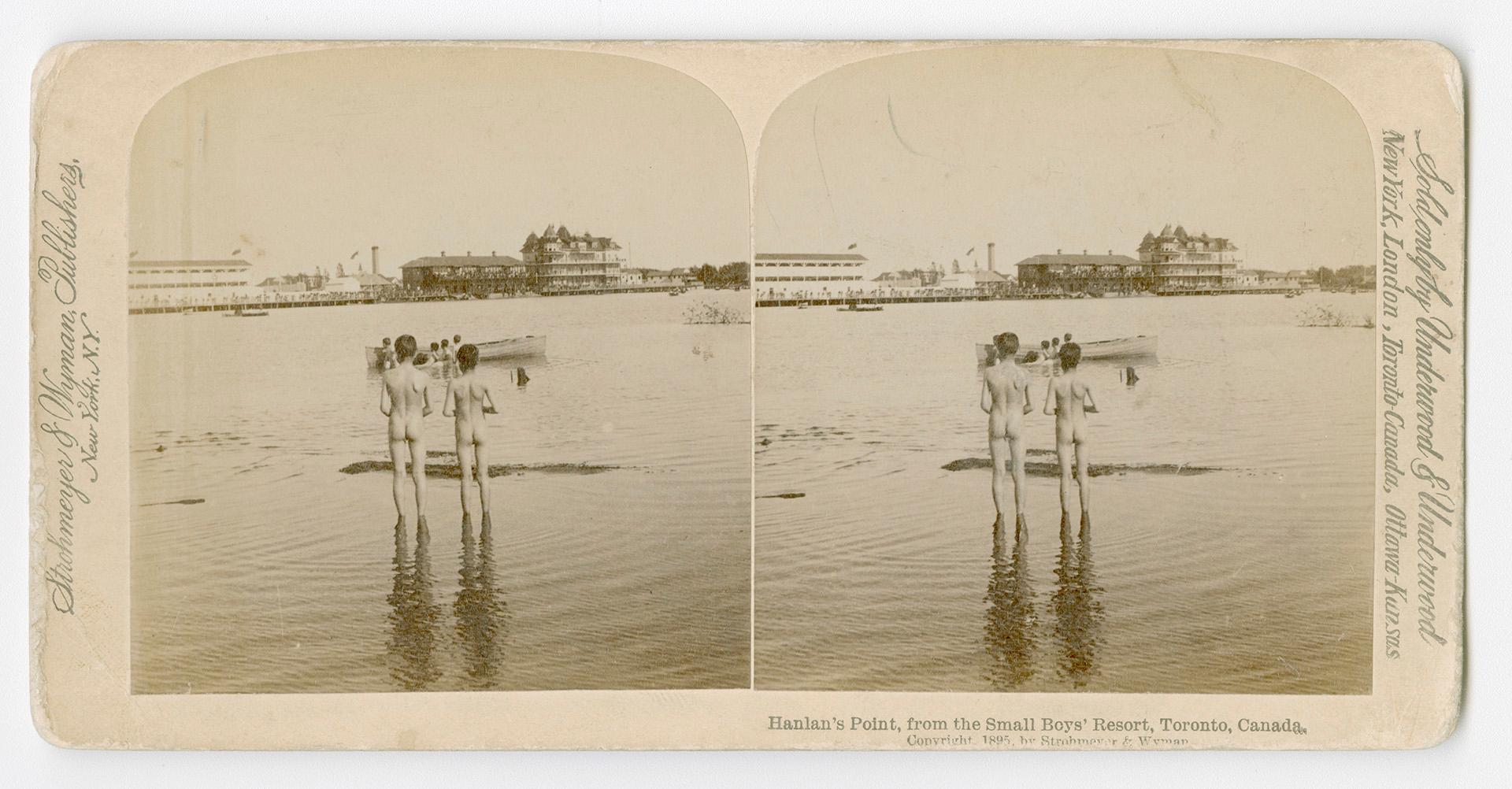 Pictures show two naked small boys watching a boat on the water with buildings on the far side.