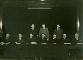 A posed photograph of ten men facing the camera, seven sitting at a long table and three standi ...