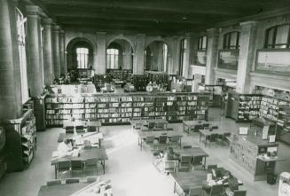 View of large library room with tables and shelves of books and arches and windows.