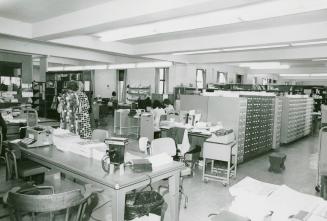 Picture of desks and card catalogues in a library work room with some staff in the background s ...