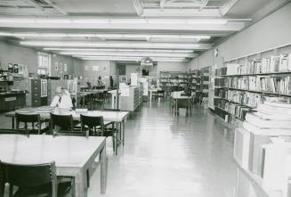 View of a library room with tables and shelves of books lining the wall and one man studying at ...