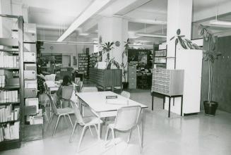 A room in a library with study tables and one person at a table, library shelves with books and…