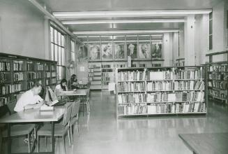 Room in a library with three tables and people studying at them and shelves of books and large  ...