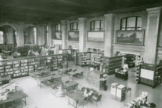 View of large library reading room with many tables, rows of shelves with books and large arche ...