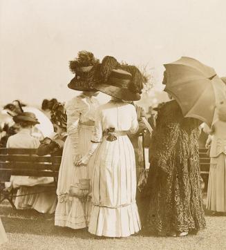 Three women wearing formal clothing and hats stand on grass in front of a bench that other simi ...
