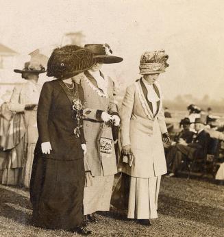 Three women wearing formal clothing and hats stand on grass in front of other, similarly dresse ...