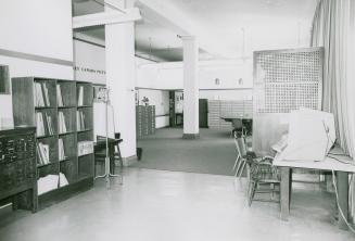 A sparsely furnished library room with tables with microfilm reading equipment, filing cabinets ...