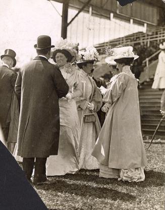 Four women and two men wearing formal clothing and hats stand on grass in front of a grandstand ...