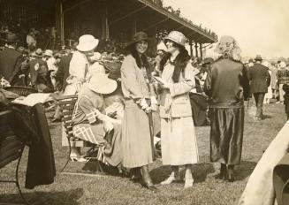 Two smiling women wearing formal dresses and hats stand in front of a large crowd of people at  ...