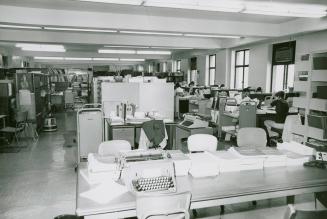A crowded library work room with a large table filled with books and a typewriter, desks, book  ...