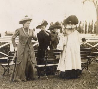Four women wearing formal dresses and hats stand around two benches in front of a racetrack. On ...