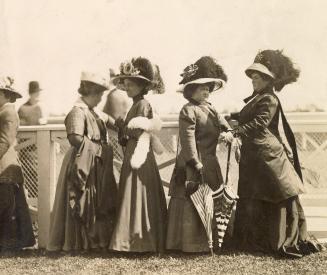 A group of five women wearing formal dresses and hats stand in front of a fence by a racetrack. ...
