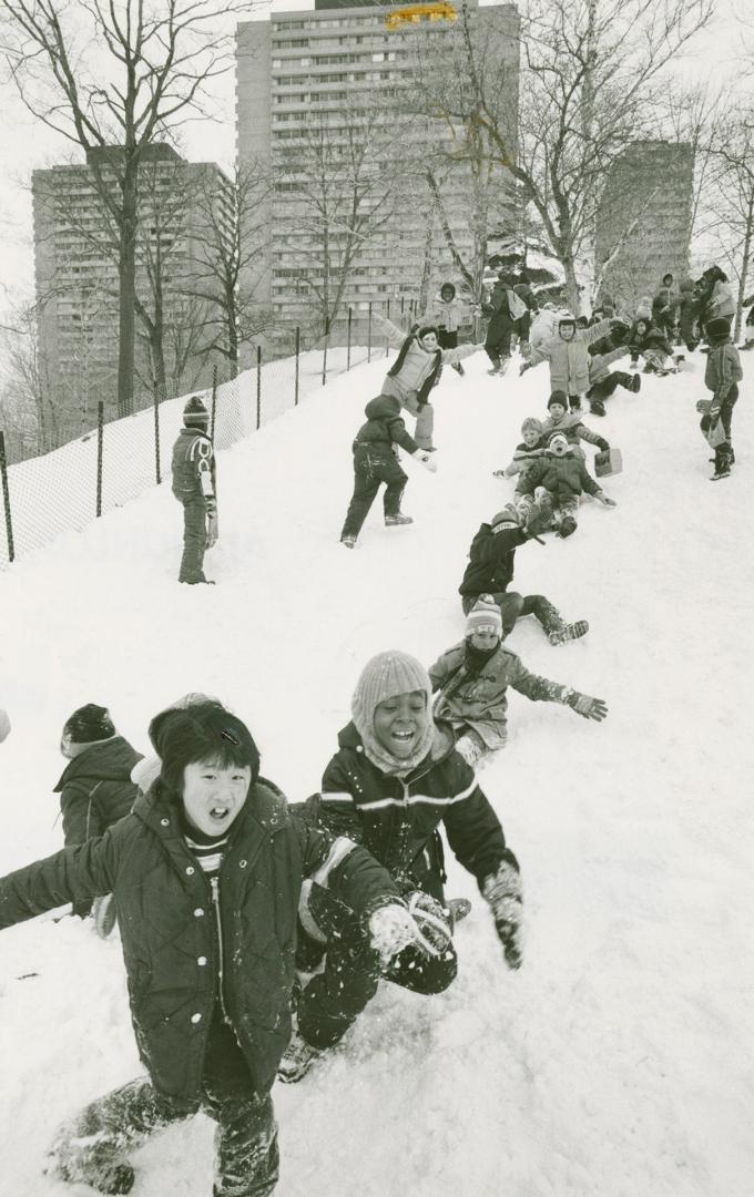 Photograph of children playing in the snow on a hill in front of apartment buildings in Fleming…