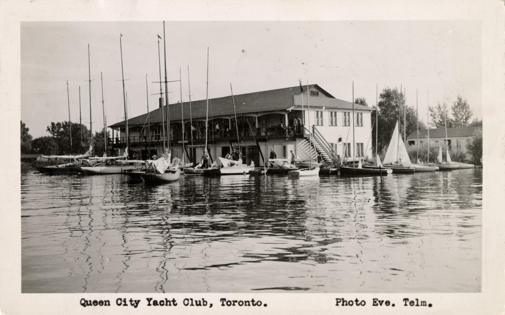 Black and white photograph of a frame building with a long porch on the side of a lake with man…