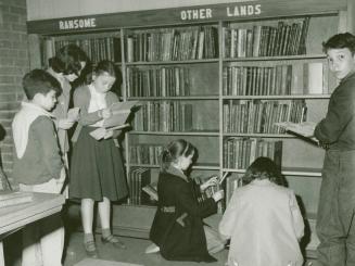 A group of children standing in front of a wall of book shelves and looking at or holding books ...