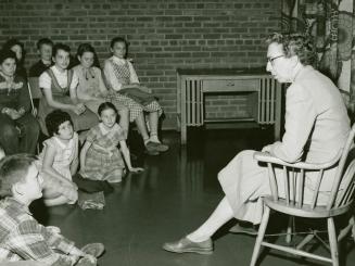 Picture of a librarian sitting in a chair reading telling a story to a group of children seated ...