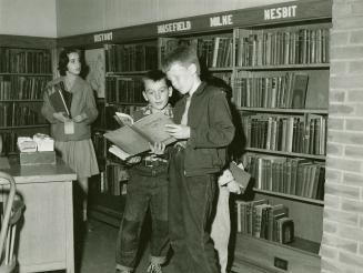 Picture of three children in a library standing looking at books from the shelves behind them. 