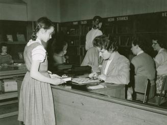 Picture of a girl in a library checking out a book at a desk with a librarian. 