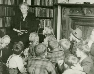 Photo of a librarian reading a book to a group of children seated around her on the floor. 