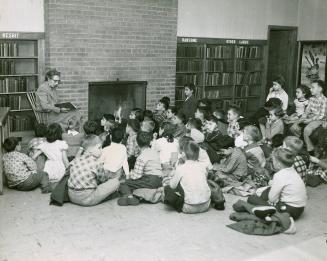 Photo of a librarian reading a story to a group of children sitting around her on the floor in  ...