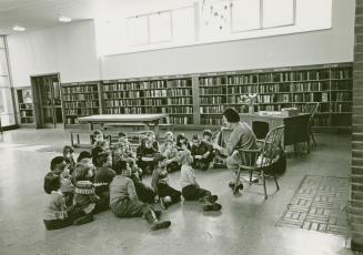 Photo of librarian reading a story to a group of children seated on the floor in a large room w ...