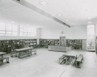 Photo of large room in a library with shelves of books lining the walls and tables and benches  ...
