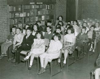 Photo of an audience of children seated in rows of chairs in room with shelves of books. 