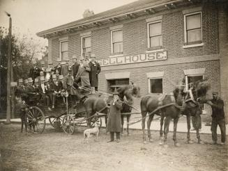 Shows a large number of men in top-hats and coats standing on an open-topped wagon, drawn by a …