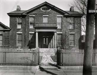 Photo of a stone house with nine sash windows and two storeys. There is a wooden picket fence a ...