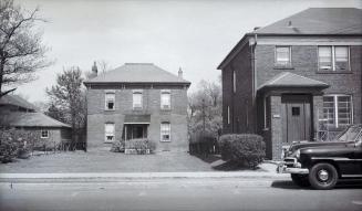 A photograph of a two-story residential house, with a road and a parked car in the foreground.  ...