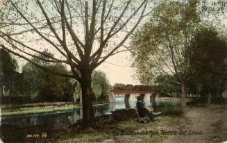 Colorized photograph of a man sitting on a park bench by the side of a river. In the background ...