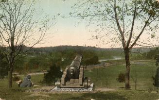 Colorized photograph of a man sitting down beside a cannon overlooking a valley with a river in…