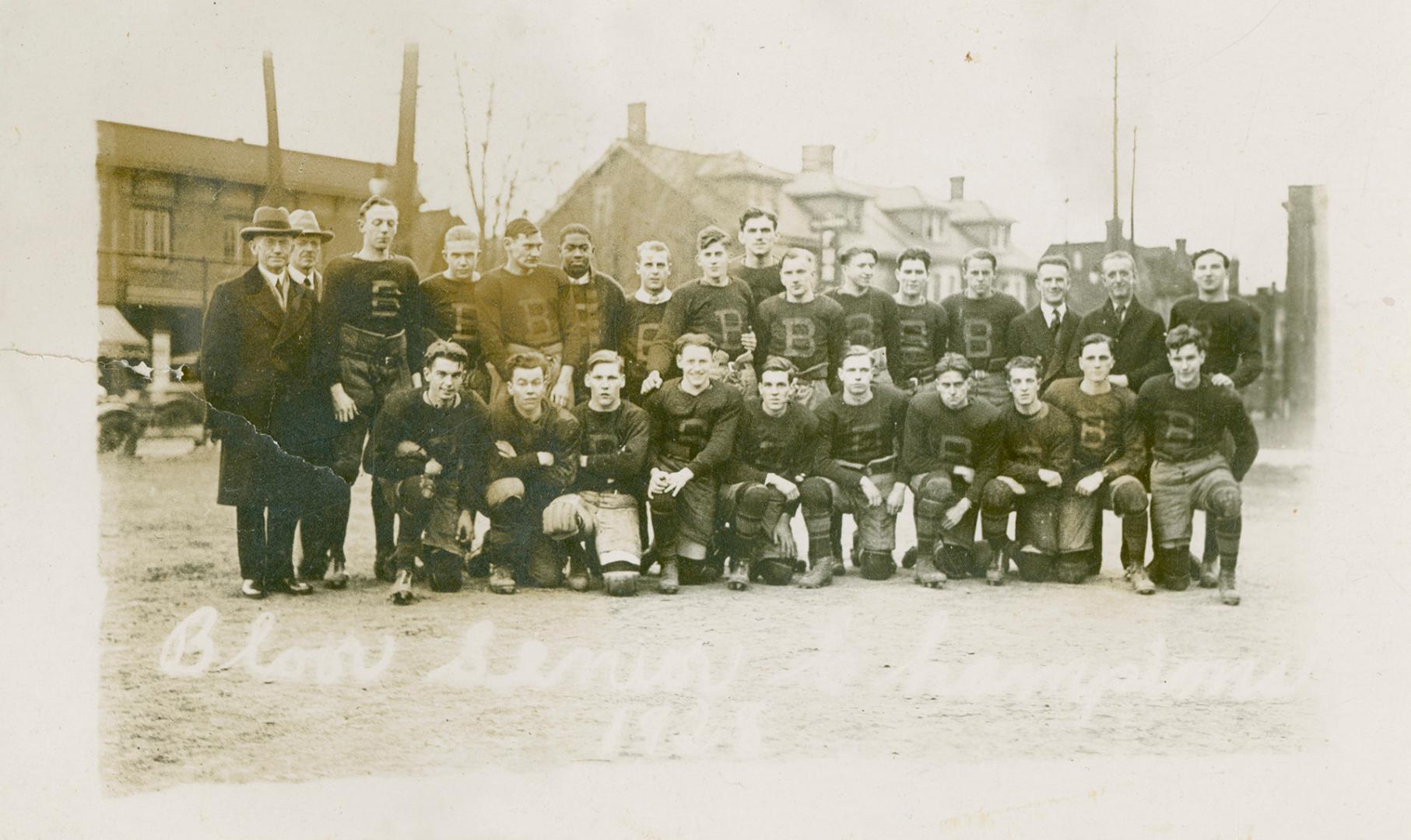 Twenty-two high school aged boys and four men pose for a team photo on the lawn of their school…