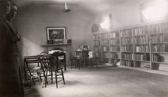 Picture of a room with tables and chairs and one wall of book shelves and tow women peaking int ...
