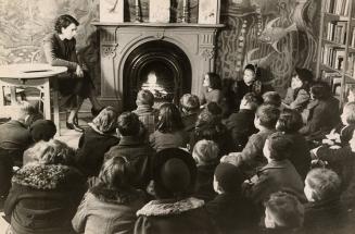 Picture of a librarian telling a story to a group of children seated on the floor gathered arou ...