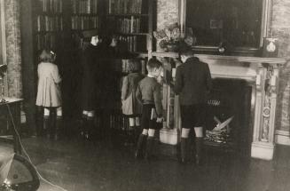 Picture of a group of children looking at library books in a room with a large fireplace and bo ...