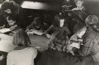 Picture of a group of children seated and standing at a table reading books in a library.
