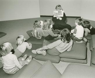 Picture of a librarian reading a book to a group of seven children seated on cushions on the fl ...