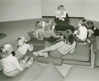 Picture of a librarian reading a book to a group of seven children seated on cushions on the fl…