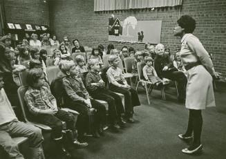 Picture of children seated in rows on chairs in a room listening to a librarian storytelling. 