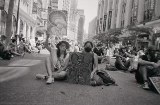 A photograph of a number of people sitting in the middle of a street during a protest. Some are ...